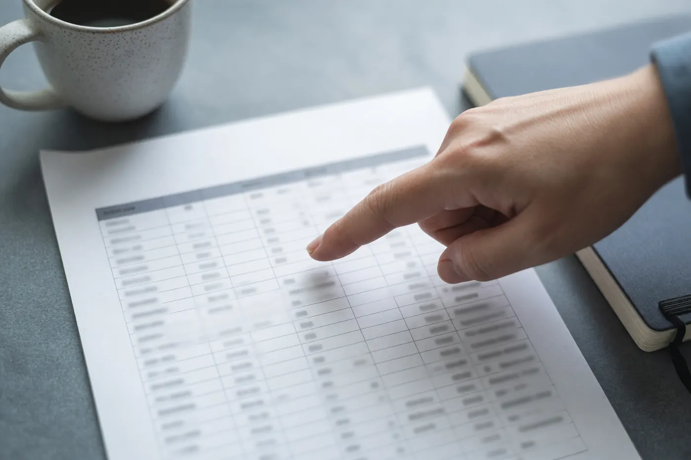 A hand pointing at a printed table of rows and columns beside a coffee mug and a closed notebook, the figures too blurred to read