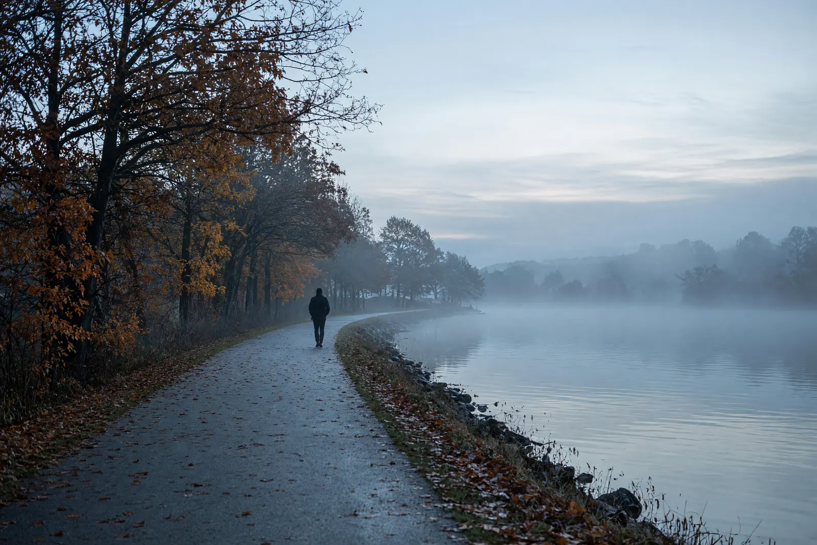 A paved lakeside path in early morning mist with autumn trees on one side and a person walking away in the distance